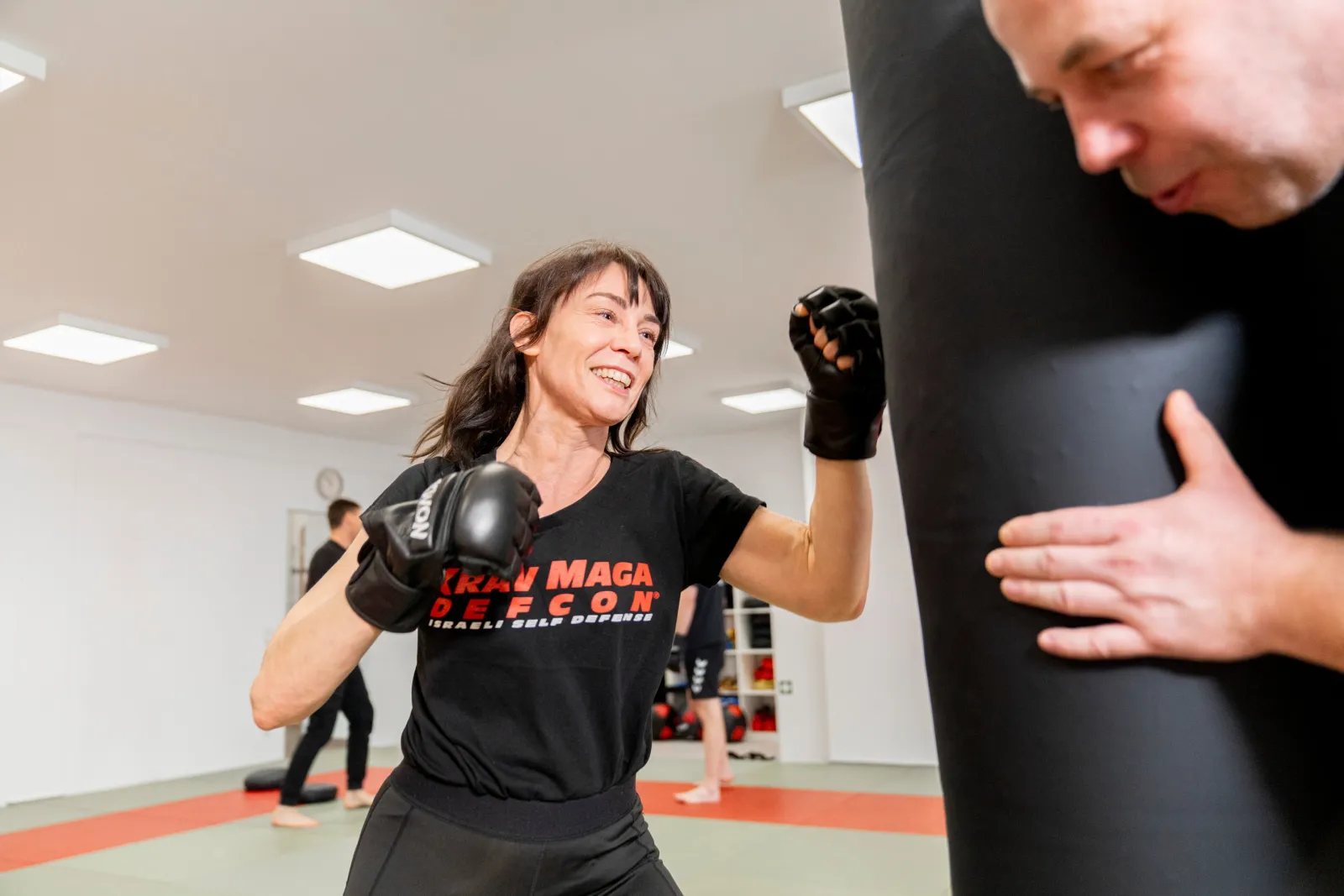 Trainierende im Krav-Maga-DEFCON-Shirt schlägt lachend mit Boxhandschuhen gegen einen Sandsack