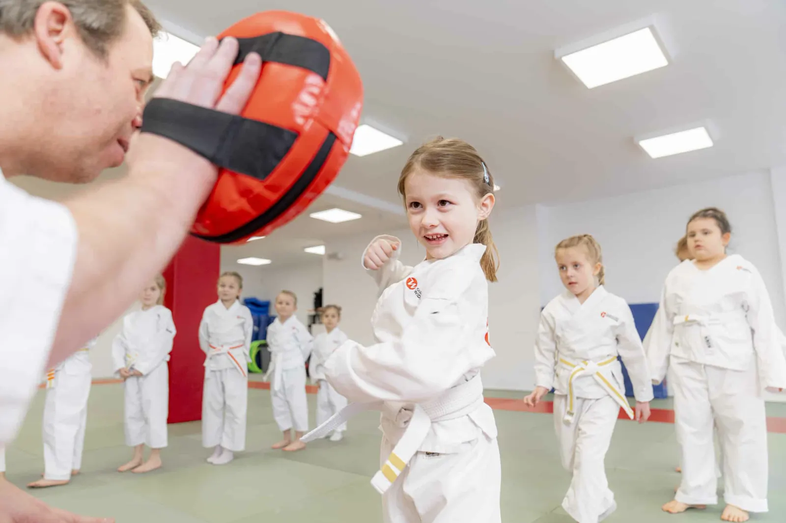 Lachendes Mädchen schlägt im Kindertraining gegen eine Pratze, andere Kinder schauen zu