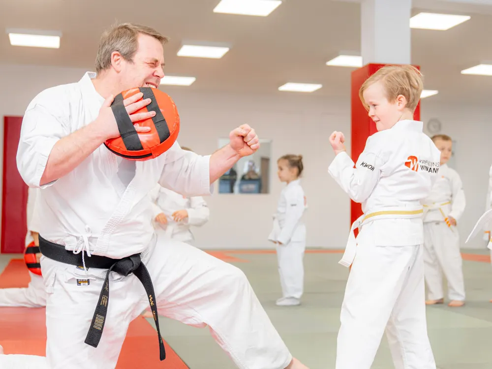 Trainer kniet mit Pratze, ein kleiner Junge holt im Kindertraining zum Schlag aus